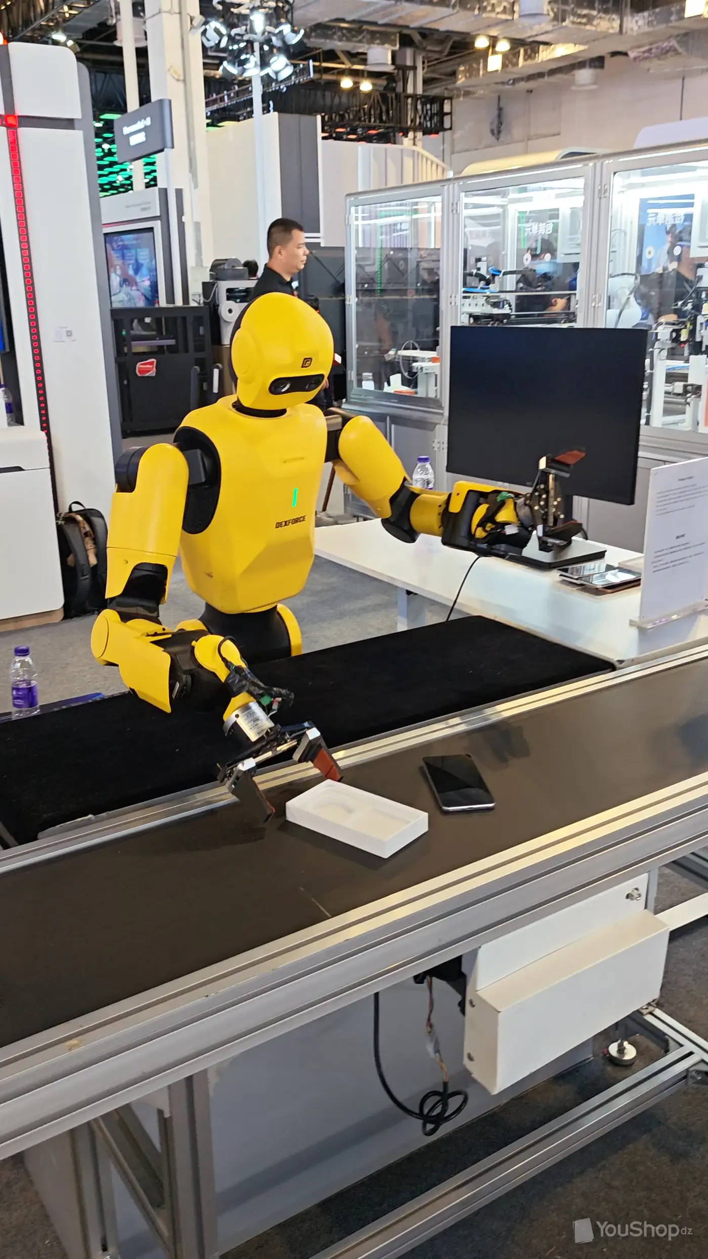 Robot humanoïde jaune DEXFORCE saisissant un objet au-dessus d’un convoyeur de démonstration